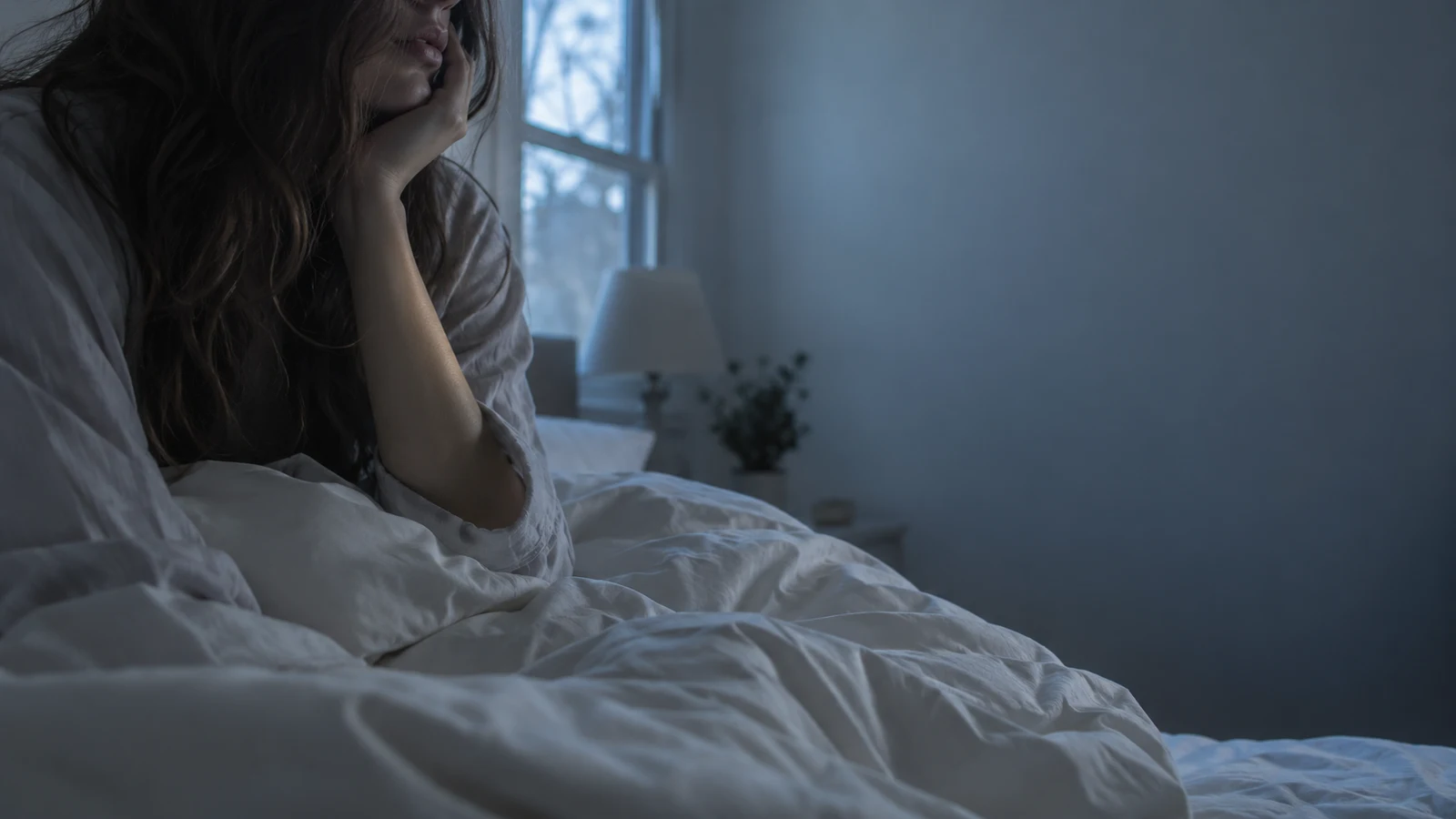 Mulher sentada na cama de manhã cedo com a mão apoiada no rosto, em quarto com luz fria