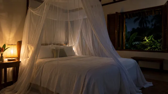 Mosquitero blanco sobre una cama tendida, con luz suave de lámpara y una ventana abierta a la noche